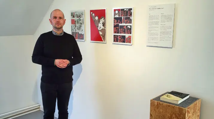 Ein Mann steht vor einer Wand an der Beispiele der Ausstellung zu sehen sind. Auf einem Sockel liegt ein Ansichtsexemplar einer Graphic Novel.